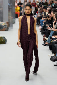 Model walks a fashion runway wearing a burgundy halter-neck jumpsuit with a deep center V, gold dotted pattern, and wide-legged pants; audience and photographers line the catwalk behind her.