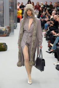 Model on a runway wearing a taupe fur-trimmed hooded coat with a long fur scarf, carrying a black structured handbag.
