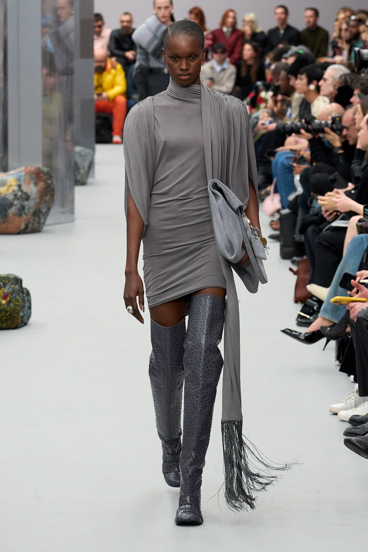Model on a runway wearing a gray draped dress with cape sleeves, gray perforated thigh-high boots, and a fringed gray handbag; audience watches in the background.