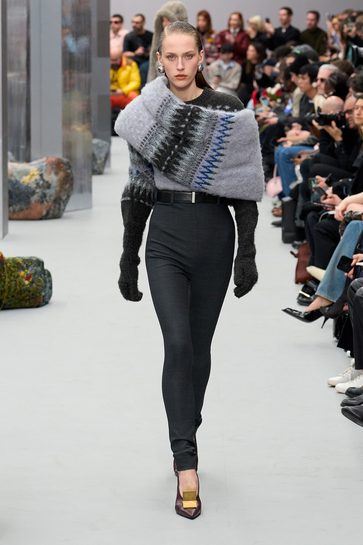 Model walks a runway wearing a chunky gray knit wrap draped over black high-waisted pants and burgundy heels with a gold buckle; audience seated along the catwalk behind her.