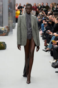 Model walks the runway in a gray plaid blazer over a dark polka-dot bodysuit, paired with burgundy heels, as photographers and spectators watch.