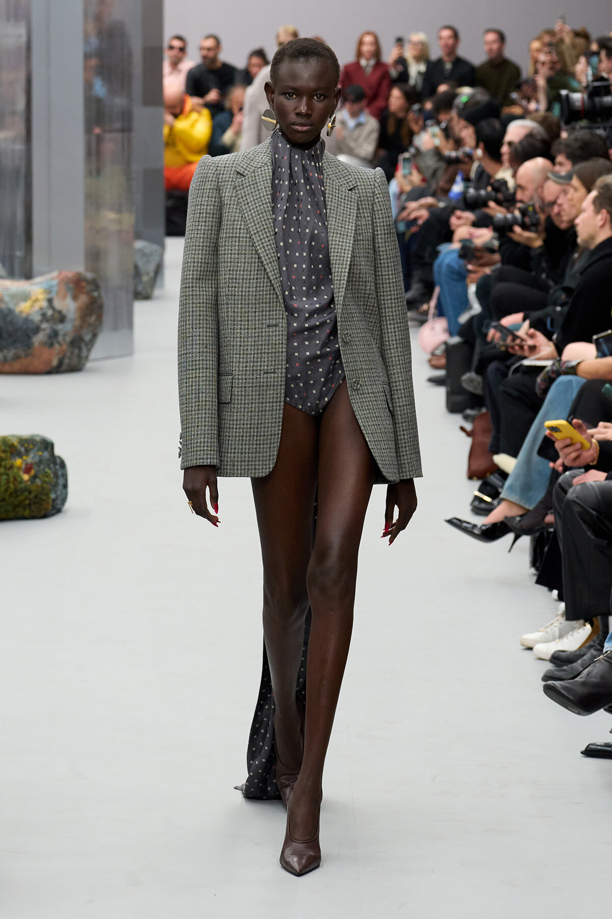Model walks the runway in a gray plaid blazer over a dark polka-dot bodysuit, paired with burgundy heels, as photographers and spectators watch.