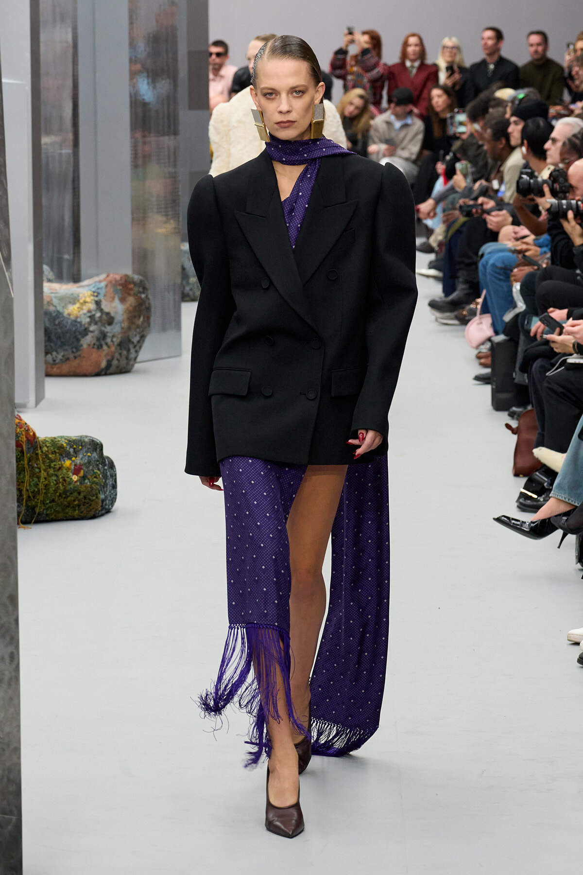 Model walks the runway in a black double‑breasted blazer over a purple fringed dress and scarf; audience watches from the sides.