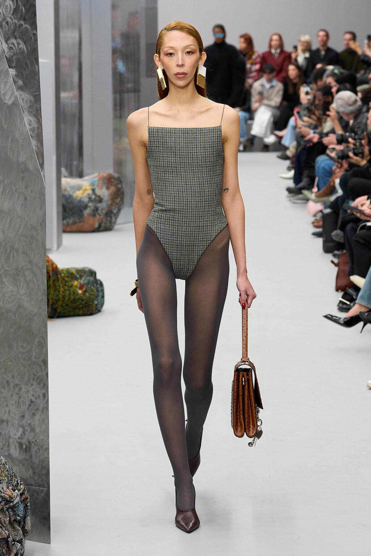 Model walks the runway in a plaid bodysuit with sheer black tights, brown heels, oversized gold earrings, and a brown structured handbag; audience watches.