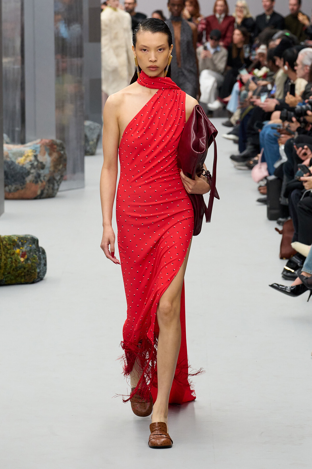 Model walks the runway in a red one-shoulder dotted dress with fringe at the hem, carrying a burgundy handbag.