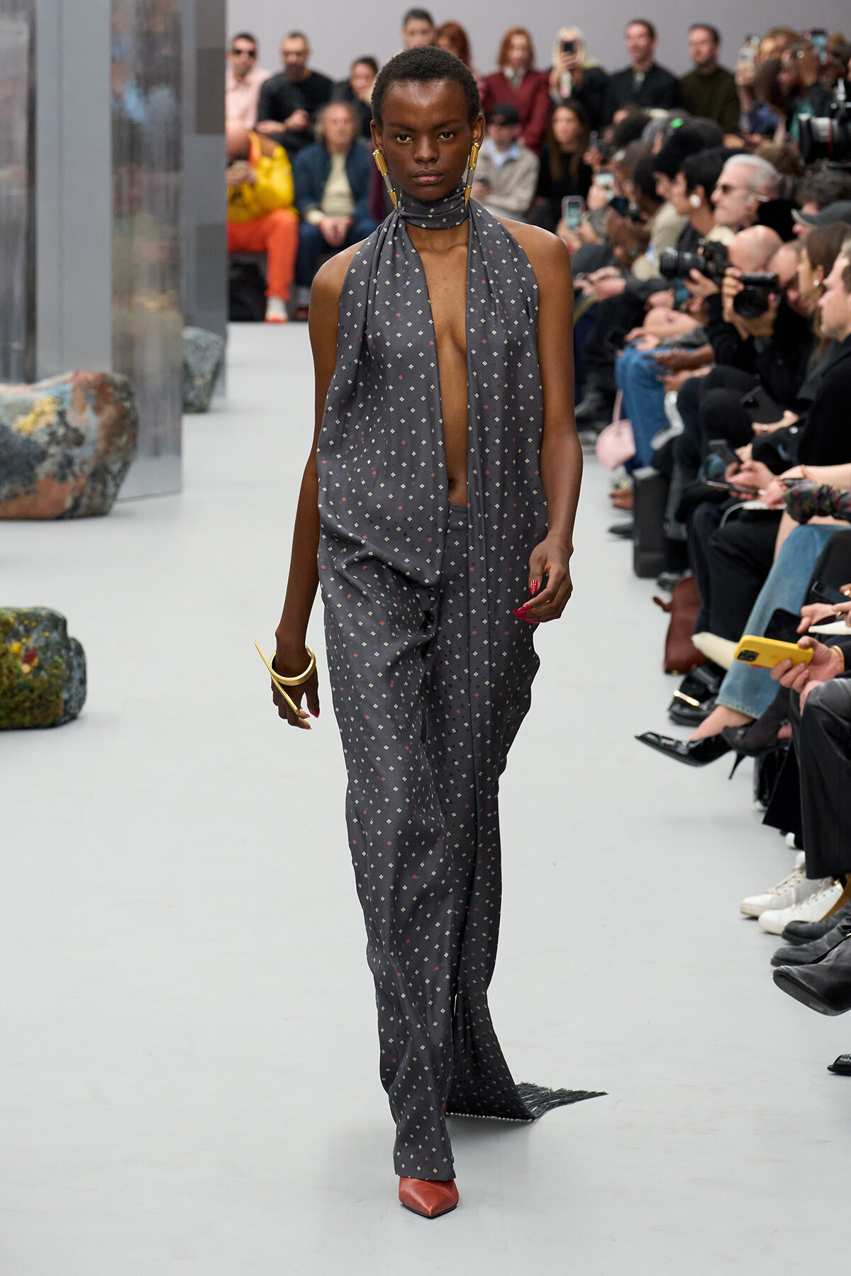 Model walking a runway in a gray dotted halter jumpsuit with a scarf, gold jewelry, and red shoes, audience in the background.