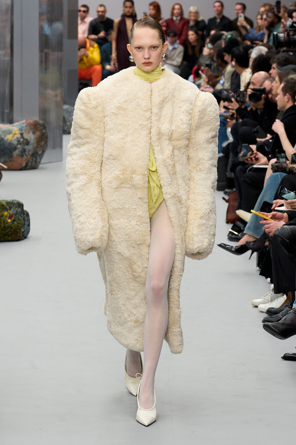 Model walks a fashion runway wearing an oversized cream shaggy coat with a lime-green dress peeking through, white heels, and a lined audience watching.