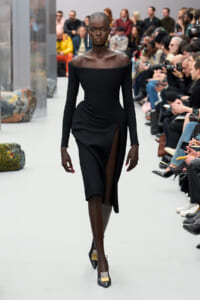 Black model walking the runway in an off-the-shoulder, long-sleeve black dress with a high slit, paired with pointed heels and gold earrings; audience in the background.