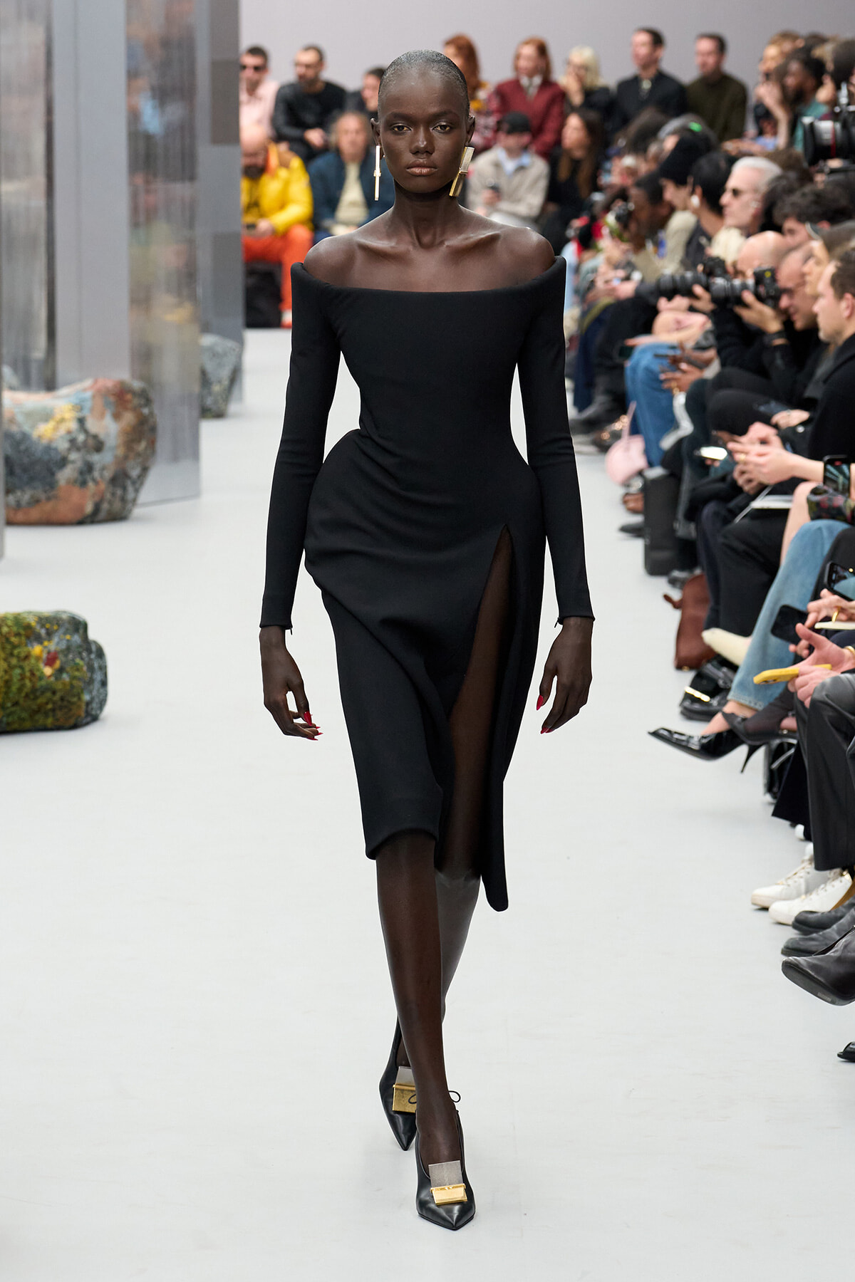 Black model walking the runway in an off-the-shoulder, long-sleeve black dress with a high slit, paired with pointed heels and gold earrings; audience in the background.