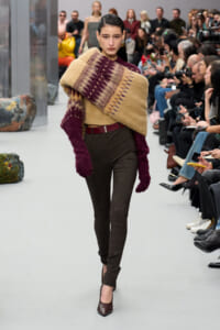 Model walking a fashion show wearing a chunky tan and burgundy knit wrap over one shoulder paired with burgundy sleeves, dark plaid pants, a burgundy belt, and burgundy heels; audience seated along the runway in the background.