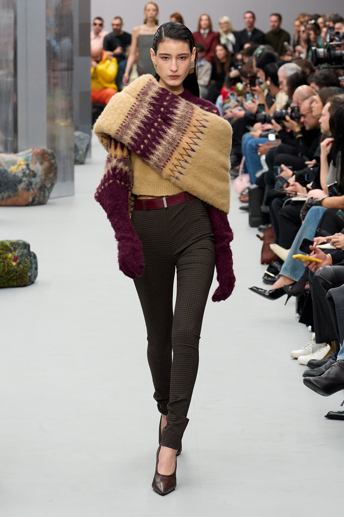 Model walking a fashion show wearing a chunky tan and burgundy knit wrap over one shoulder paired with burgundy sleeves, dark plaid pants, a burgundy belt, and burgundy heels; audience seated along the runway in the background.