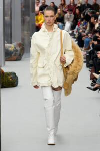 Runway model wearing a cream oversized jacket with white textured pants, white boots, and a tan fur coat draped over her shoulder with a tan bag beside it.