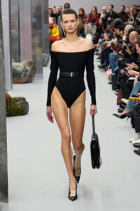 Model walking a fashion runway in an off-shoulder black bodysuit with a black belt, carrying a black handbag and wearing black heels with gold accents, audience in background