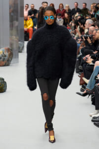 Model walks the runway in a bulky black fuzzy sweater, black knee-hole leggings, blue sunglasses, and burgundy heels with gold buckles, with an audience in the background.