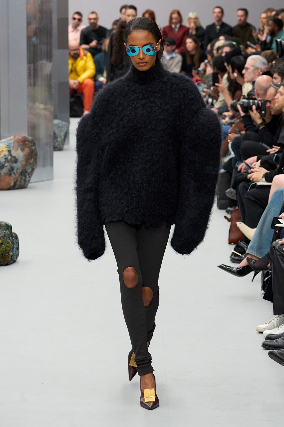 Model walks the runway in a bulky black fuzzy sweater, black knee-hole leggings, blue sunglasses, and burgundy heels with gold buckles, with an audience in the background.