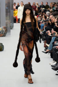 Model walks the runway in a strapless dark dress with a brown feathered/animal print pattern and long furry hooded scarf ending in pom-poms; audience watches nearby.