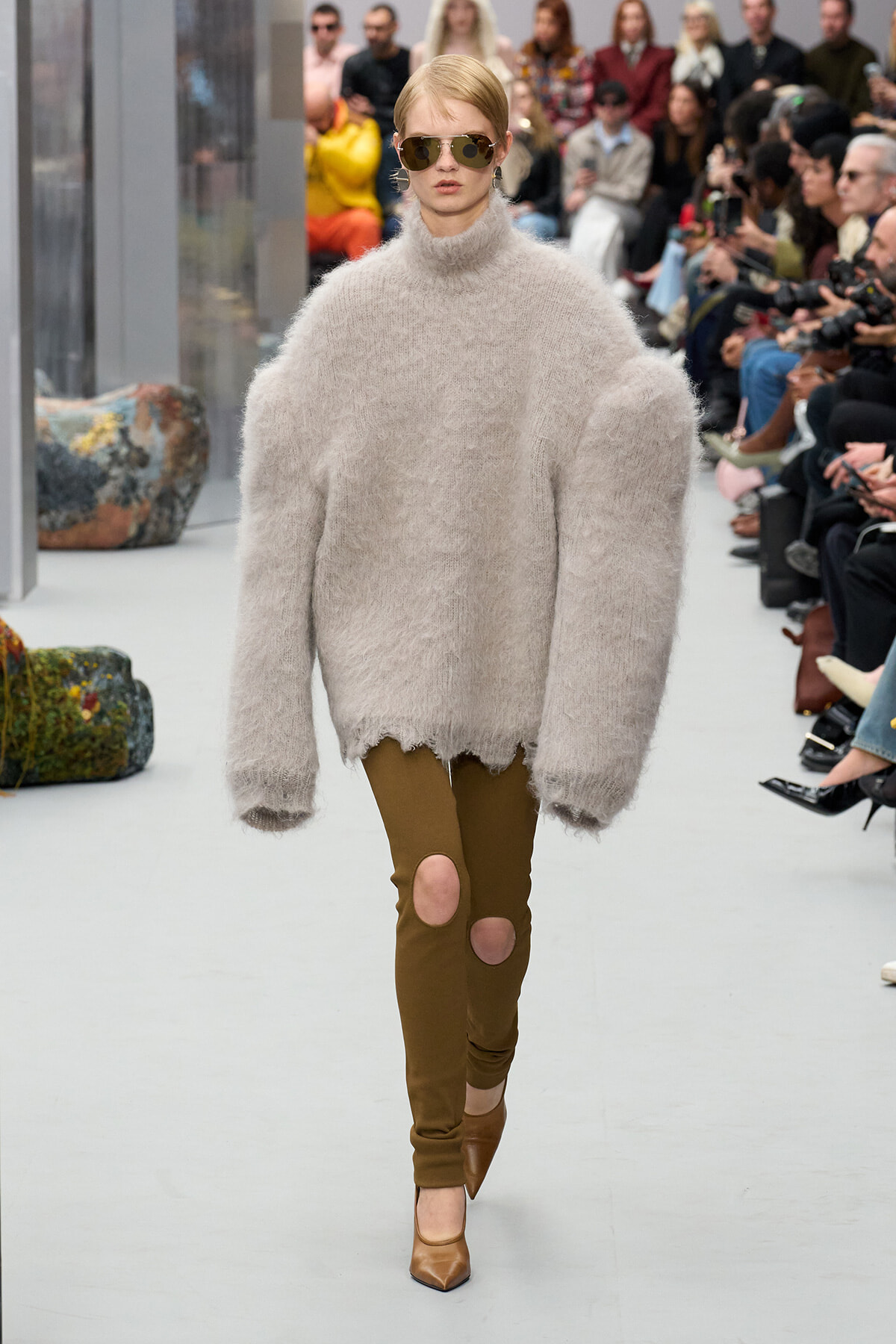 Model walking a runway in a fluffy beige turtleneck sweater, brown ripped leggings, brown heels, and sunglasses.