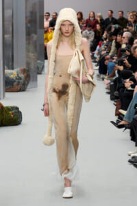Runway model in a beige strapless dress with a cream fuzzy hood and matching beige handbag, walking the catwalk.