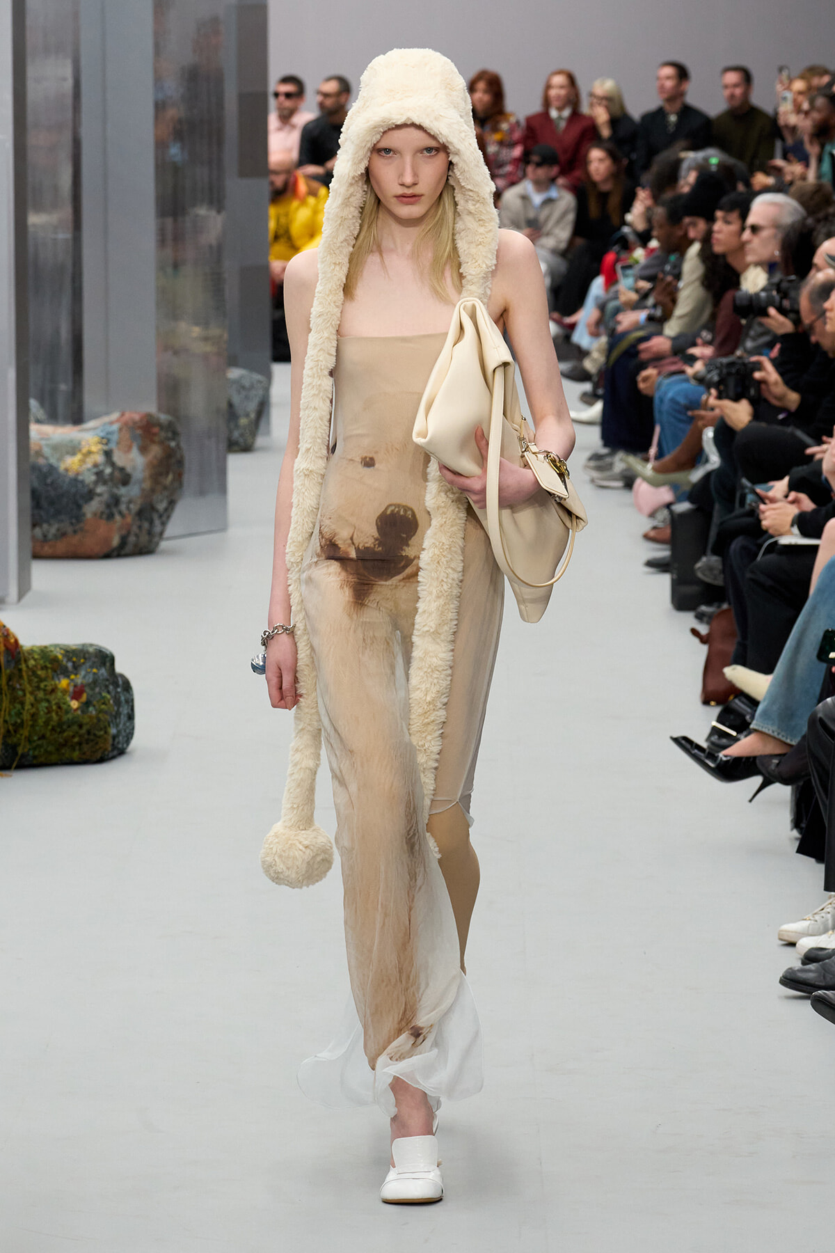 Runway model in a beige strapless dress with a cream fuzzy hood and matching beige handbag, walking the catwalk.