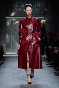 Model walks a fashion runway in a glossy burgundy snakeskin coat with belt and black heels.