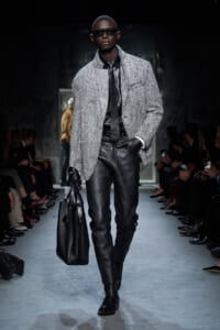 Male model on a runway wearing a textured gray overcoat, black leather pants, a black shirt and tie, gloves, and dark sunglasses, carrying a large black bag; audience along the catwalk in dim lighting.