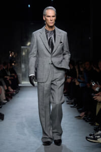Male model walking a fashion show in a gray tailored suit with a black satin shirt and gloves on the runway