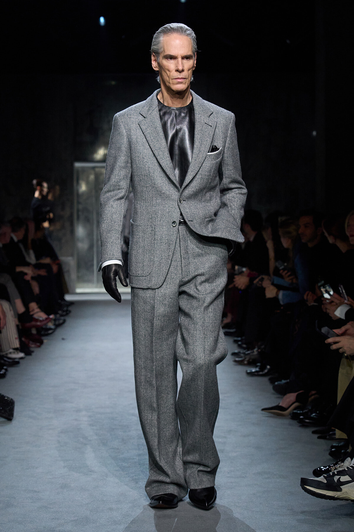 Male model walking a fashion show in a gray tailored suit with a black satin shirt and gloves on the runway