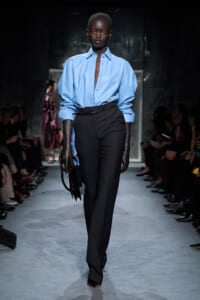 Model on a runway wearing a light blue oversized shirt tucked into high-waisted black trousers, brown belt, and dark gloves while carrying a structured dark handbag.
