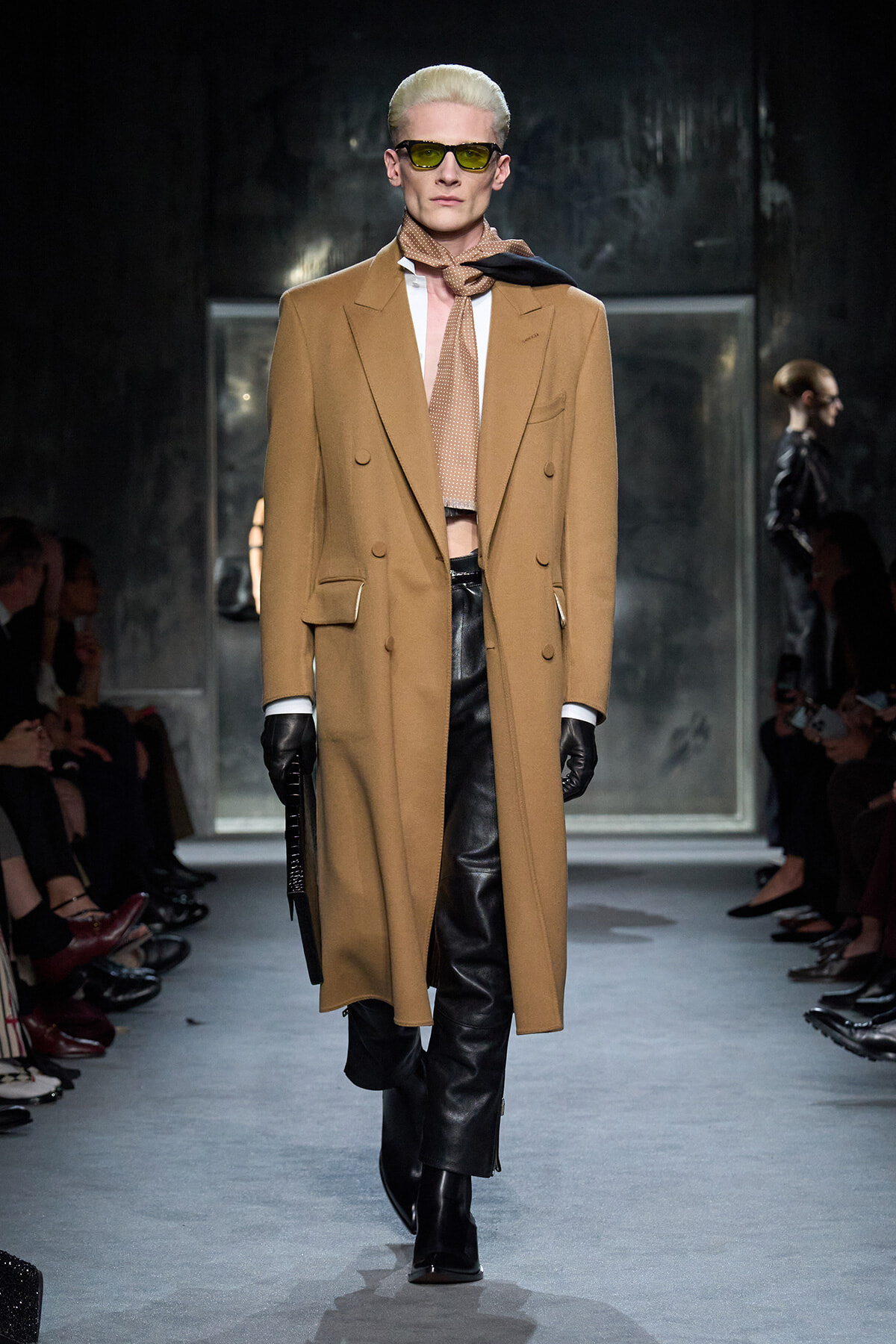 Model walking the runway in a camel double-breasted coat, black gloves, and black leather pants with a beige scarf and yellow sunglasses.