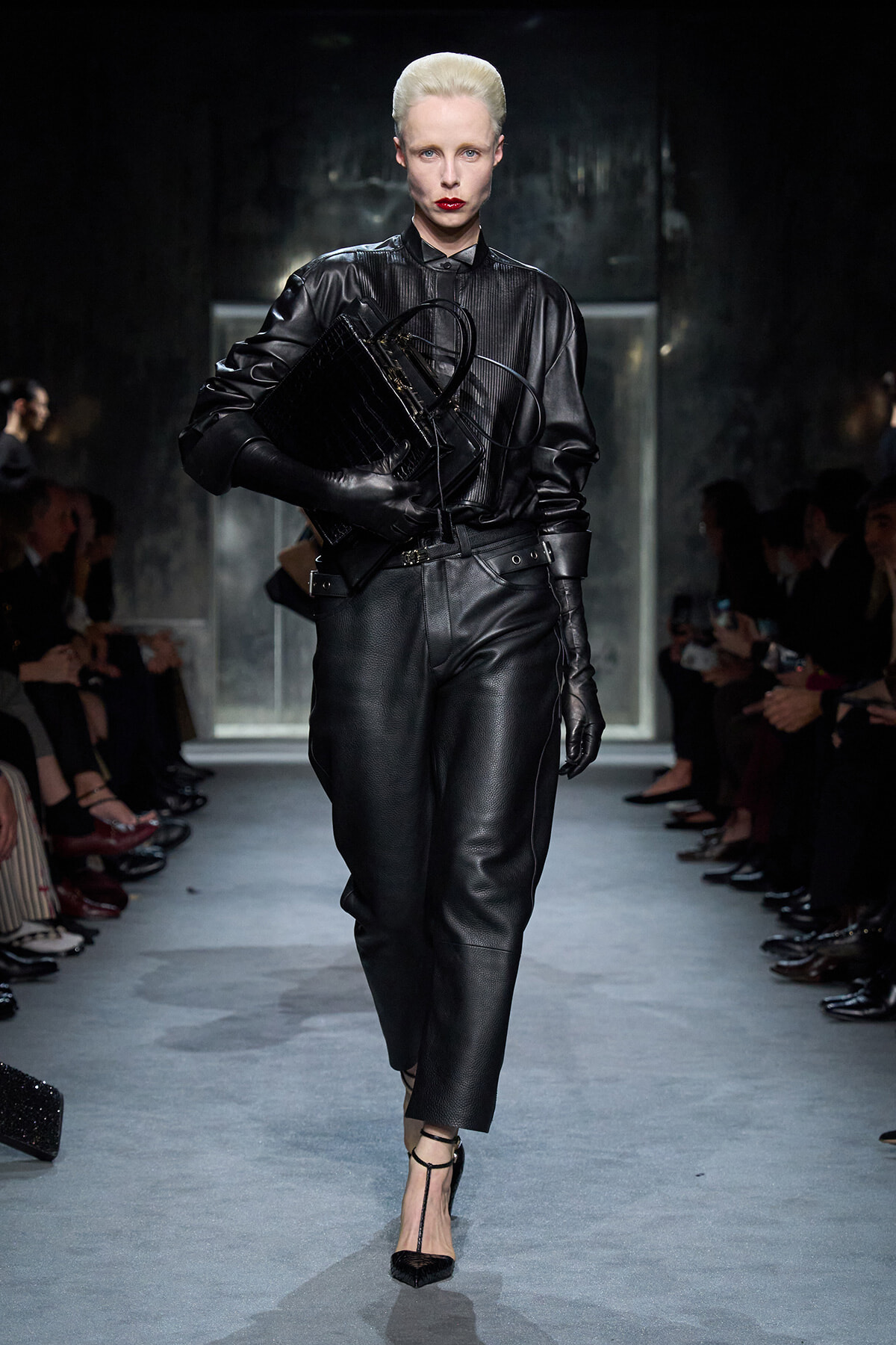 Model on a fashion runway in a black leather ensemble, holding a large black handbag; audience seated along the sides.