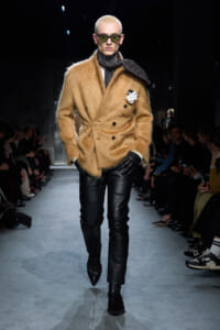 Model on a runway wearing a tan fuzzy double-breasted coat, black leather pants, gloves, scarf, and sunglasses with a white flower accessory on the lapel.