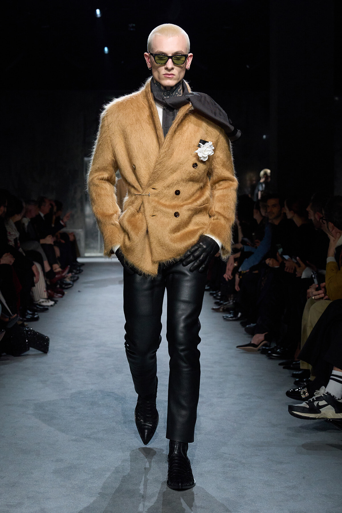 Model on a runway wearing a tan fuzzy double-breasted coat, black leather pants, gloves, scarf, and sunglasses with a white flower accessory on the lapel.
