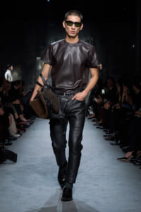 Male model walking a runway in a dark leather outfit with sunglasses, carrying a fur-lined brown bag.