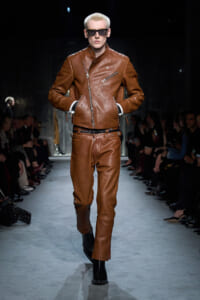 Model on a runway wearing a brown leather biker jacket and matching pants, hands in pockets and wearing sunglasses.