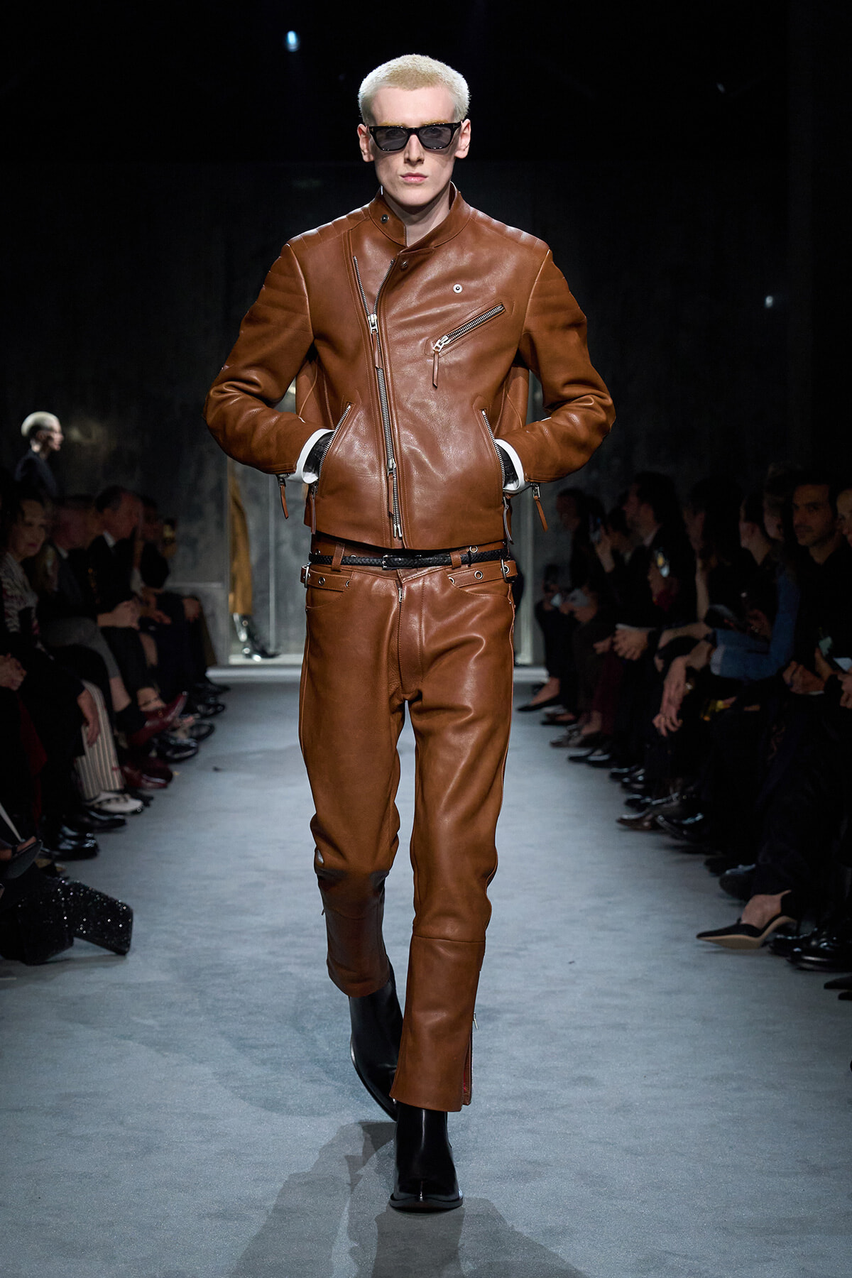 Model on a runway wearing a brown leather biker jacket and matching pants, hands in pockets and wearing sunglasses.