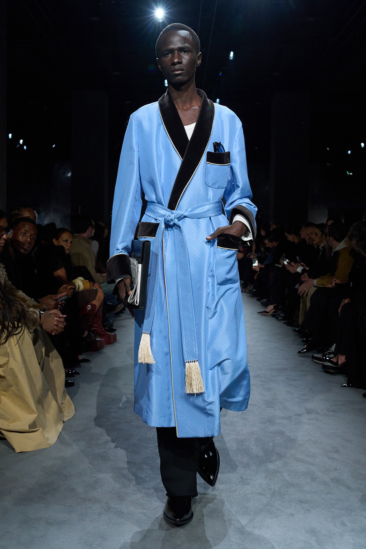 Male model on a runway wearing a light blue robe with black trim, belt, and tassels, carrying a small clutch.