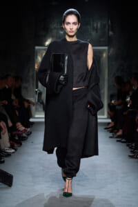 Model walking a fashion runway in an all-black ensemble: long coat, cropped top, loose pants, and green-heeled shoes, holding a black handbag and wearing gloves