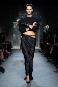 Model on a runway wearing a cropped black sweater and satin wrap skirt with bold green eye makeup and stacked cuffs on her left wrist.