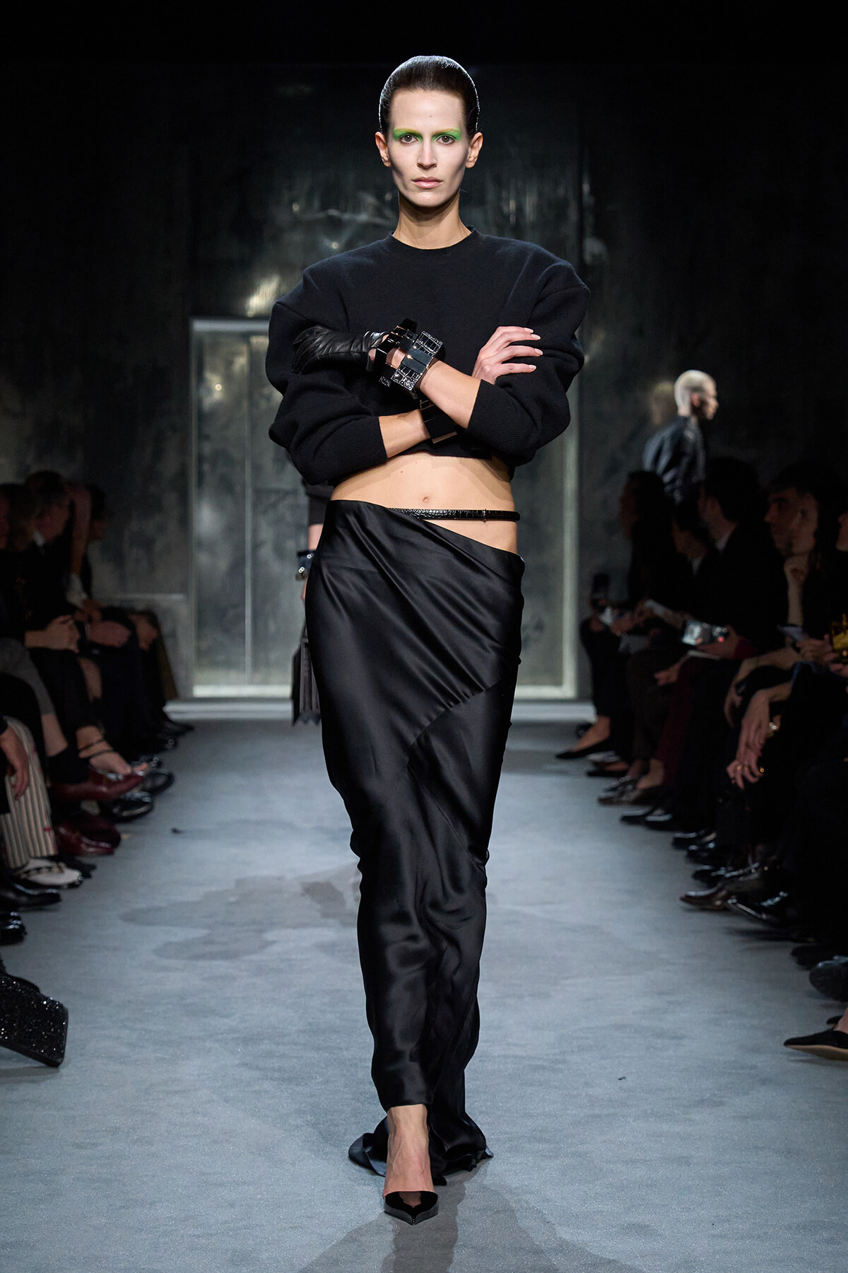 Model on a runway wearing a cropped black sweater and satin wrap skirt with bold green eye makeup and stacked cuffs on her left wrist.