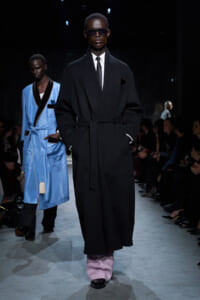 Male model on runway wearing a long black belted coat, white shirt, polka-dot tie, and lavender trousers; audience in background.