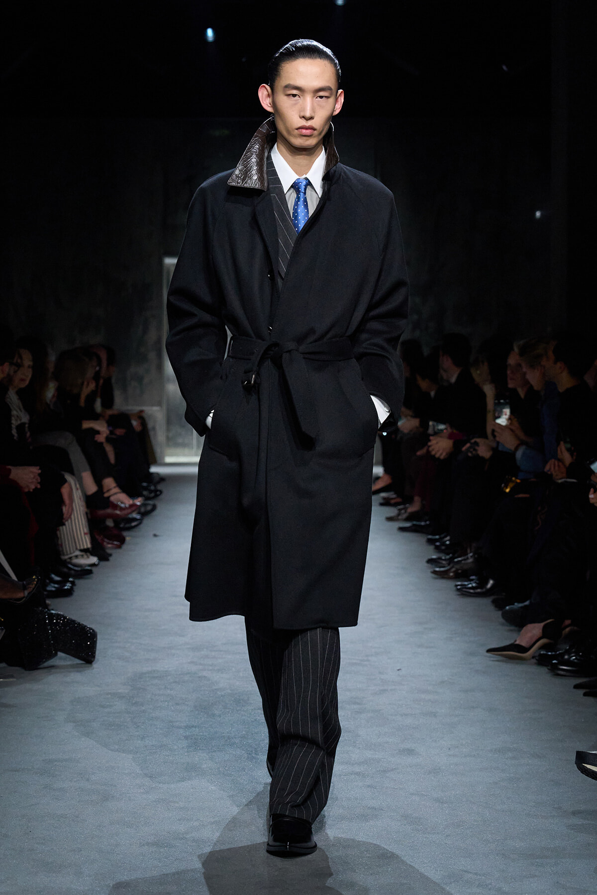 Male model walks the runway in a black belted overcoat with a brown collar, white shirt, blue dotted tie, and pinstriped trousers, audience flanking the catwalk.