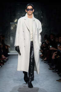 Male model on a runway wearing a long white double-breasted coat, cream sweater, and black leather pants with gloves and sunglasses.