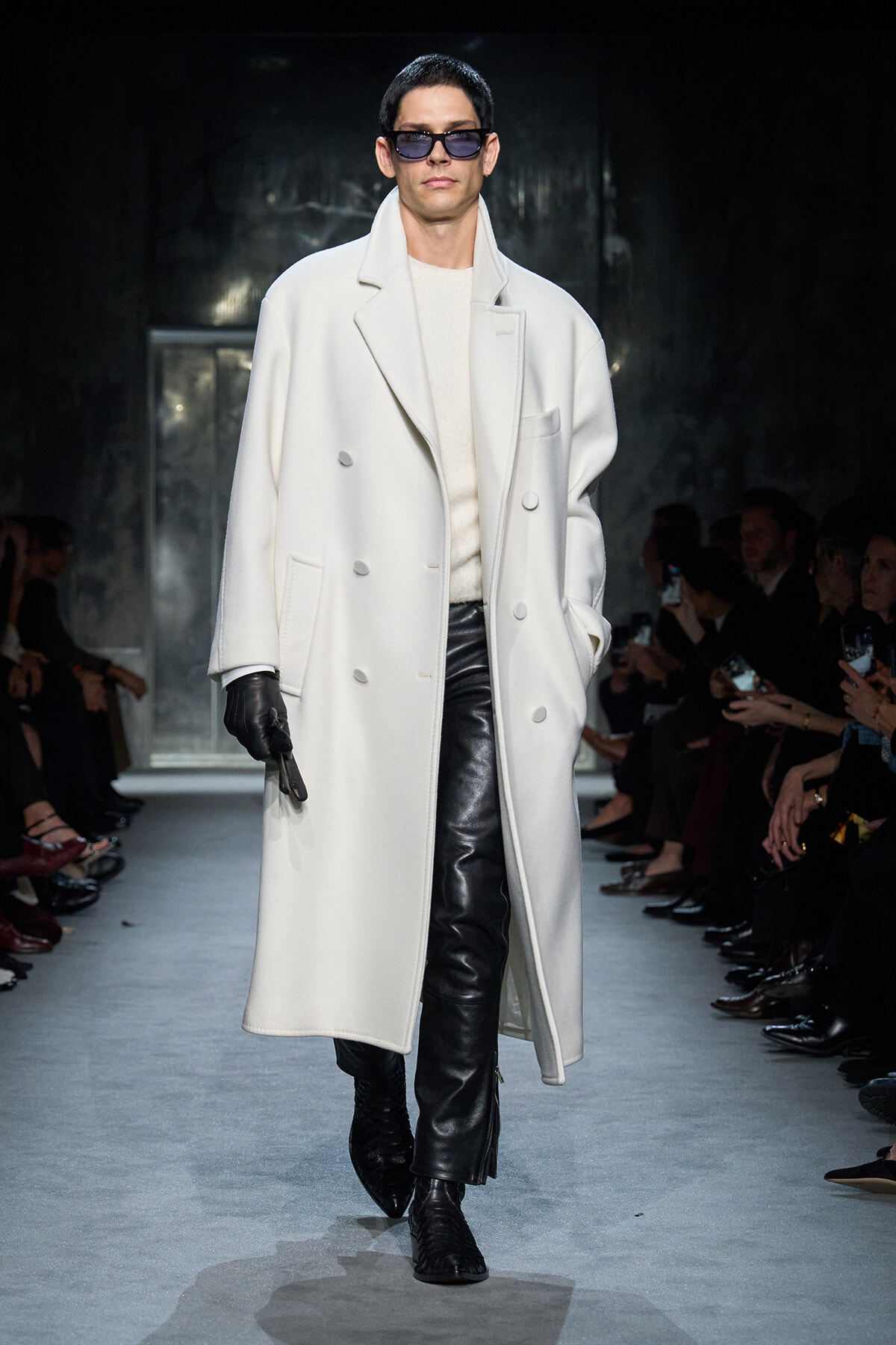 Male model on a runway wearing a long white double-breasted coat, cream sweater, and black leather pants with gloves and sunglasses.