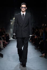 Male model on a runway wearing a black double-breasted suit, white shirt, black tie, gloves, and sunglasses.