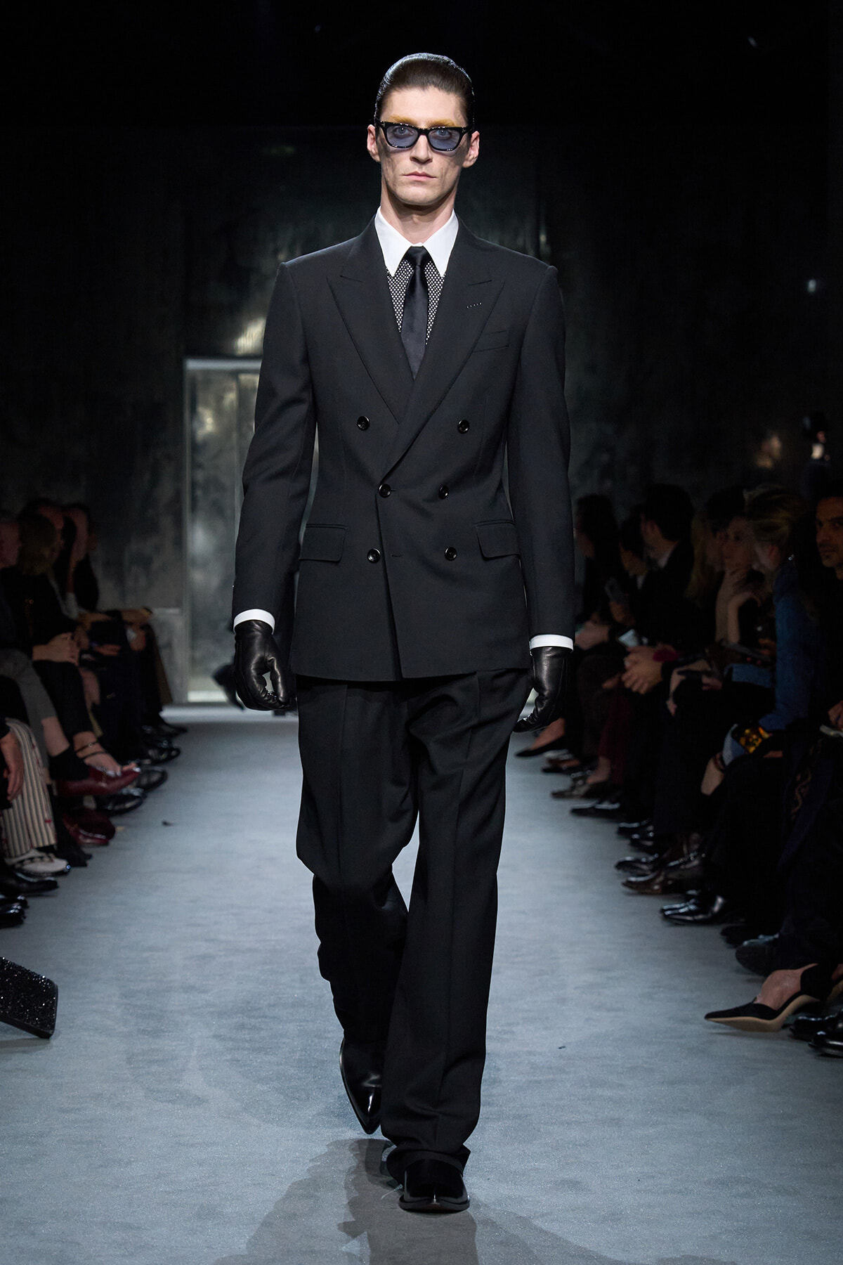 Male model on a runway wearing a black double-breasted suit, white shirt, black tie, gloves, and sunglasses.