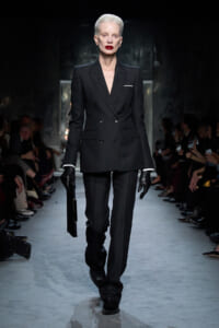 Model walking a runway in a tailored black double-breasted suit with a white pocket square, gloves, and a black clutch.