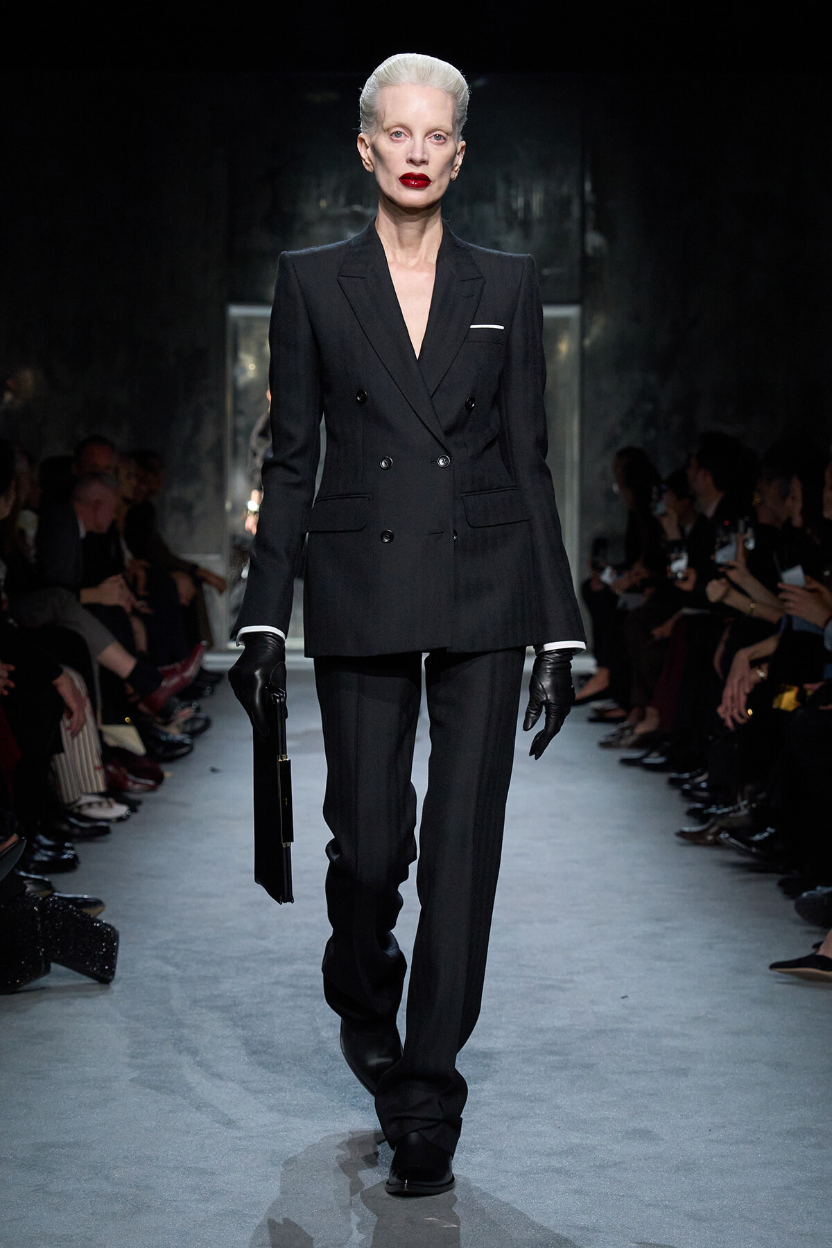 Model walking a runway in a tailored black double-breasted suit with a white pocket square, gloves, and a black clutch.