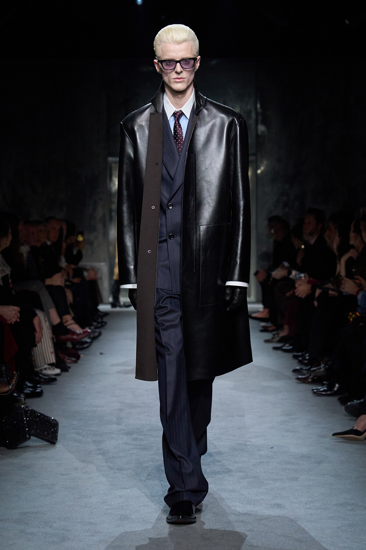 Male model on runway wearing a long black leather coat over a dark pinstripe suit and blue tie, with blonde hair and glasses.