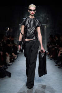Model walks the runway in a black leather top, black wide-leg pants, and oversized dark sunglasses, carrying a black coat.