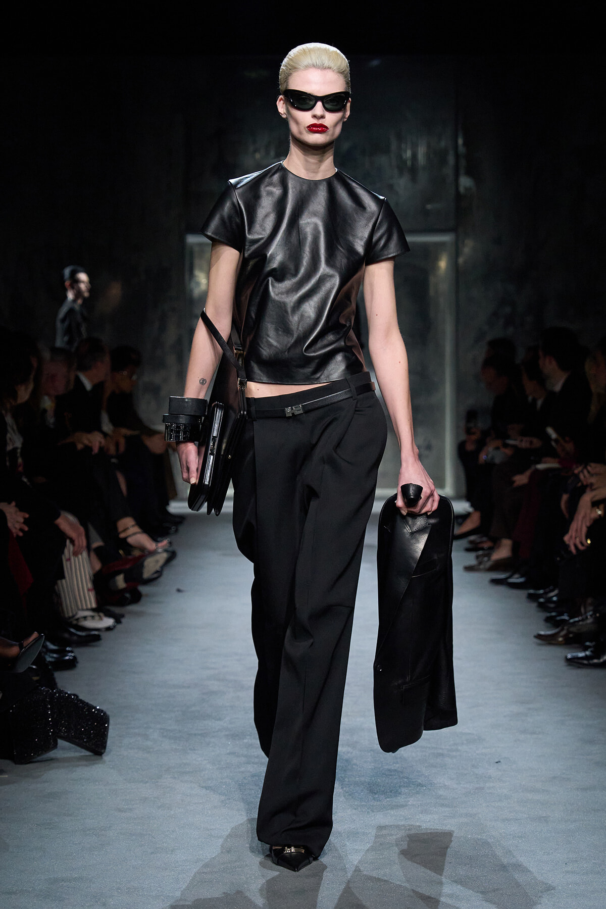 Model walks the runway in a black leather top, black wide-leg pants, and oversized dark sunglasses, carrying a black coat.